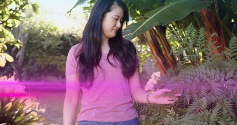Young woman inspecting fern in tropical backyard, wearing pink tee and jeans