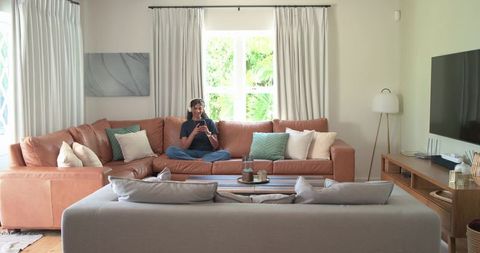 African american woman sitting on sectional using headphones and smartphone in cozy living room
