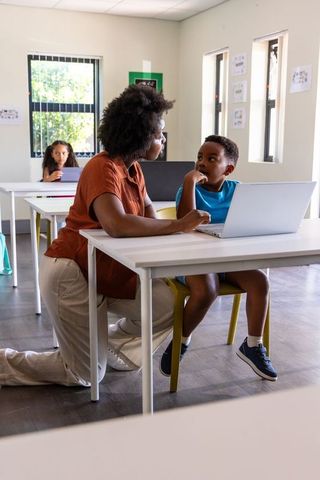 Teacher Kneeling Helping Student Using Laptop in Modern Classroom During Digital Learning