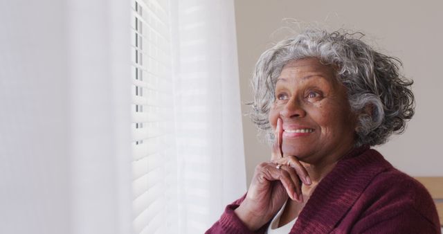 Elderly African American woman with grey hair smiling while looking out a window. Bathed in natural light with white curtains and blinds, the scene captures a peaceful, content, and thoughtful moment. Ideal for use in ads or articles about aging gracefully, elder care, happiness in later life, or everyday life of seniors.