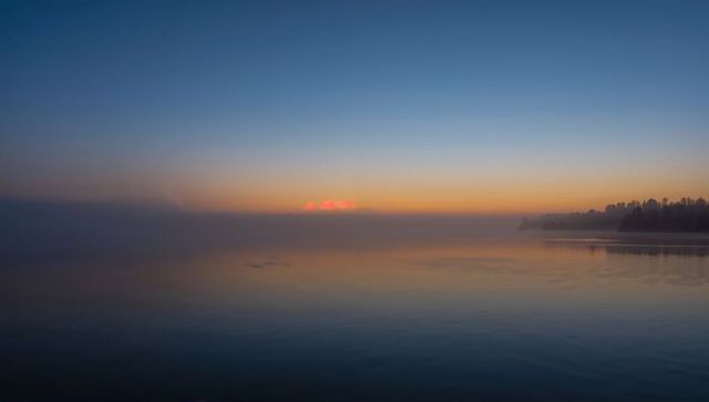 Misty Dawn Over Tranquil Lake Reflecting Sunrise Hues and Silhouetted Shoreline