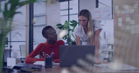 Collaborating businesswomen leaning over laptop in glass office workspace project planning