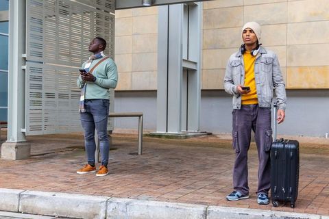 Two commuters waiting at bus shelter checking phones with rolling suitcase and scarves