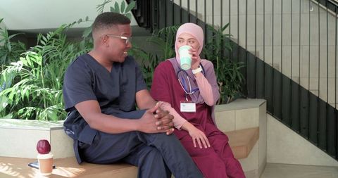 Black nurse and hijab-wearing colleague sharing coffee and conversation in hospital lobby