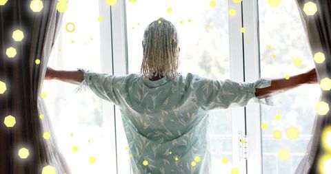 Senior woman opening curtains to sunlit balcony morning wearing light-green botanical robe