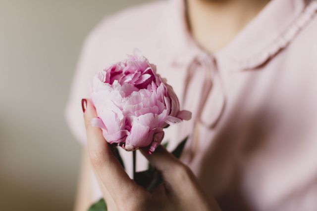 Woman holds pink peony flower in hand, highlighting delicacy and beauty of both the flower and femininity. Can be used in themes related to nature, romance, gardening, beauty, femininity, and peaceful moments.