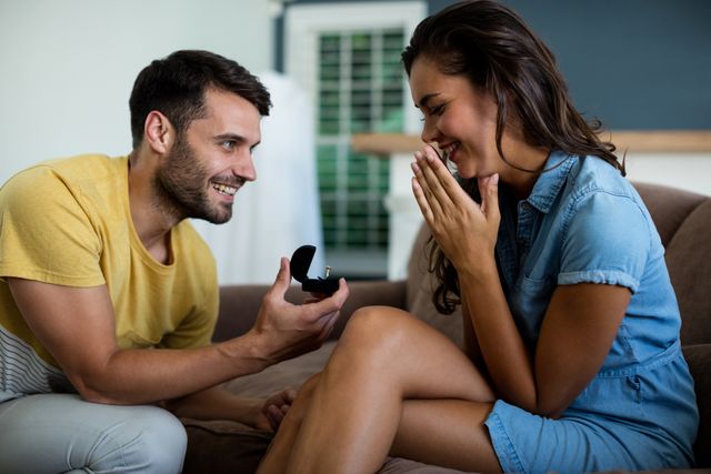 Man proposing to woman with engagement ring in living room. Woman appears surprised and happy. Perfect for use in articles, blogs, and advertisements related to engagements, relationships, and romantic moments.