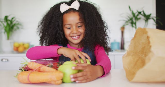 Young girl with curly hair and pink shirt unpacking groceries, including apples and carrots, in bright kitchen. Ideal for themes related to healthy living, family lifestyle, child nutrition, and home activities.