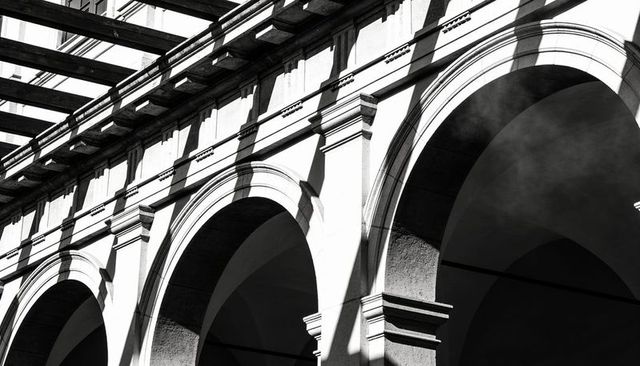 Monochrome sunlight casting diagonal shadows across classical arches and colonnade