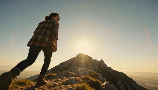 Woman hiking ridge toward mountain peak at golden hour with lens flare and backlit silhouette
