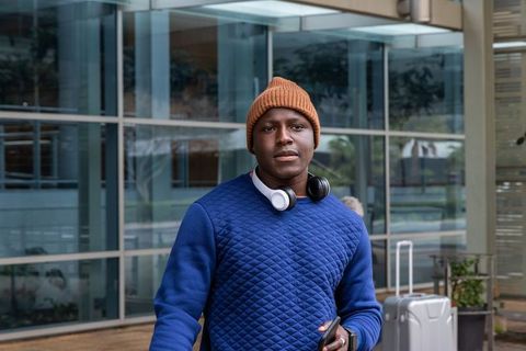 Young african american traveler wearing headphones and beanie with suitcase at glass entrance
