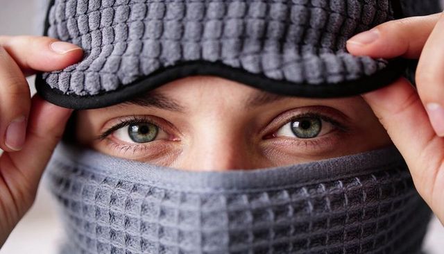 Revealing green eyes peeking through grey waffle gaiter while adjusting sleep mask