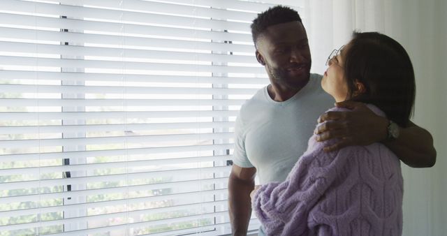 Happy diverse couple embracing and looking through window in bedroom. Spending quality time at home concept.