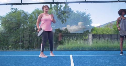 African american women playing pickleball on blue court with glass wall and green backdrop