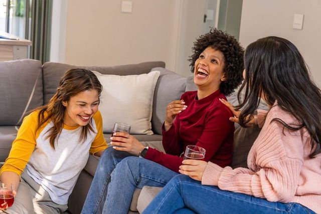 Multicultural friends laughing on gray sofa holding red drinks, casual home gathering