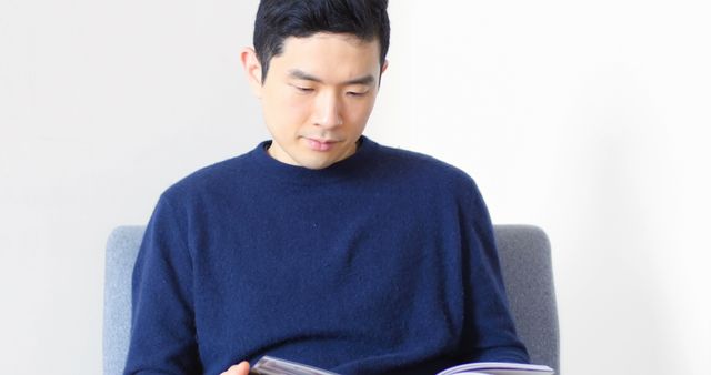 An Asian man is seated comfortably while engrossed in reading a book, with copy space. His focused expression and casual attire suggest a moment of leisure or self-education.