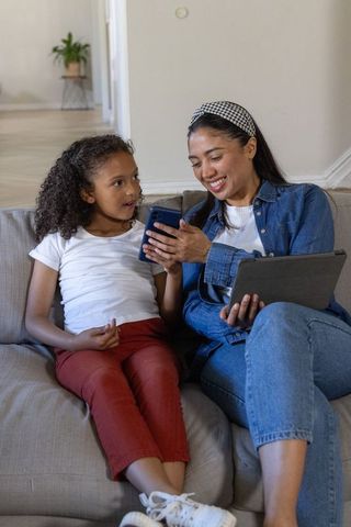 Smiling diverse caregiver showing smartphone to curious child on couch, using tablet