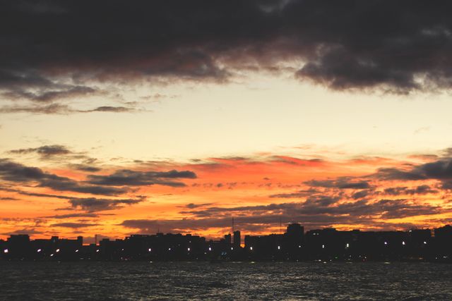 City skyline silhouetted against vibrant sunset with clouds over ocean water. Ideal for backgrounds, travel blogs, urban lifestyle promotions, or relaxation and tranquility themed projects.