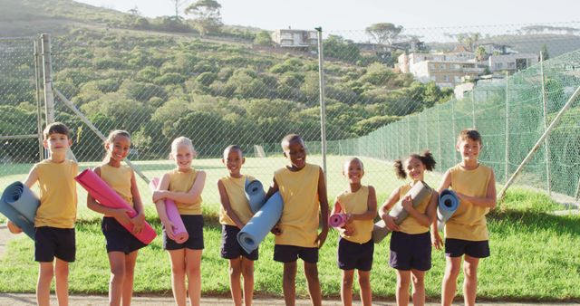 Diverse group of smiling children standing in a line, holding yoga mats outdoors in the bright sunlight. The background features lush greenery, hills, and a chain-link fence. Shows physical fitness, outdoor activities, and a sense of togetherness. Ideal for use in educational materials, fitness programs for kids, and advertisements promoting outdoor activities.