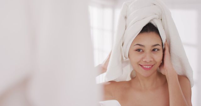 Woman wrapping her wet hair with a bath towel while smiling at a mirror. Perfect for illustrating routines related to personal hygiene, post-bath relaxation, and skincare. Can be used in advertisements for beauty products, bathroom accessories, or wellness blogs.