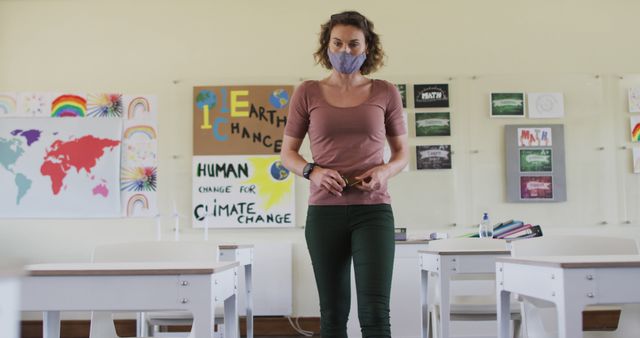Female teacher wearing mask walking through classroom with environmental posters on walls. Suggestive of educational settings, safety measures during pandemic, focusing on raising awareness about climate change and environmental protection. Ideal for themes related to education, sustainability, health, and safety protocols in schools.