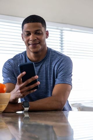 Young man checking smartphone at kitchen counter wearing smartwatch and casual blue shirt
