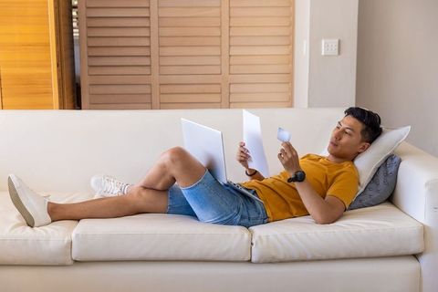 Asian man reclining on sofa, working with laptop and papers in sunlit cozy living room