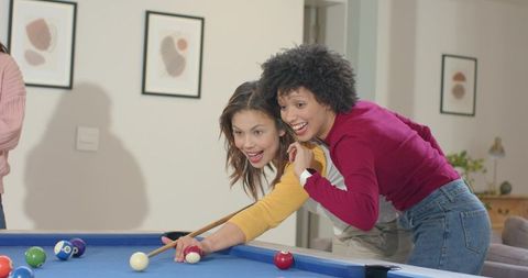 Multicultural friends playing pool at home laughing and guiding cue shot on blue felt