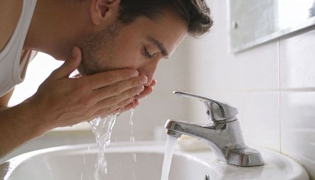 Young man washing face at bathroom sink rinsing under running faucet fresh morning routine