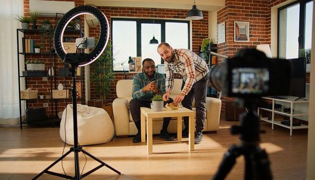 Filming Duo Wearing Plaid Shirts Collaborating on Sofa with Ring Light in Loft Studio