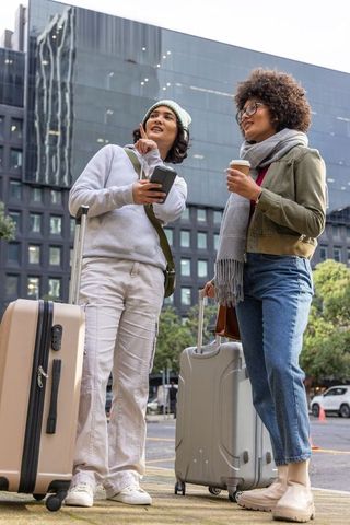 Multicultural friends navigating city streets with suitcases and coffee near glass tower
