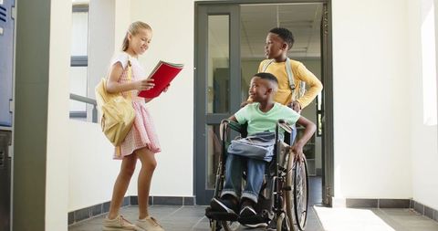 Students interacting in school corridor with wheelchair student reading red notebook