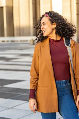 Young woman walking and smiling on sunlit urban plaza wearing blazer and shoulder bag
