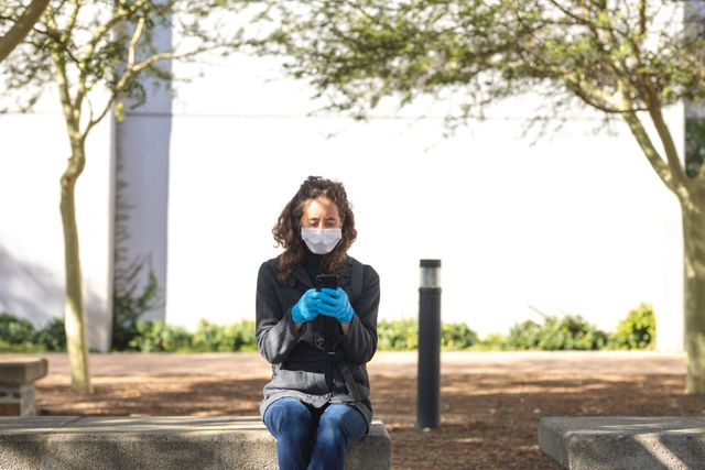 Caucasian woman out and about in the city streets during the day, wearing a face mask and gloves against coronavirus, covid 19, sitting on wall using smartphone.