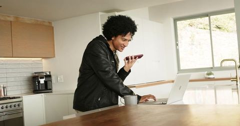 African American man speaking into smartphone while using laptop on modern kitchen island