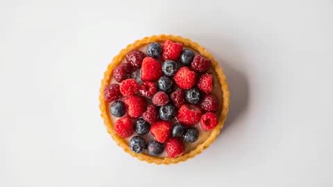 Camera panning over mixed berry custard tart on white table, overhead reveal of glossy berries