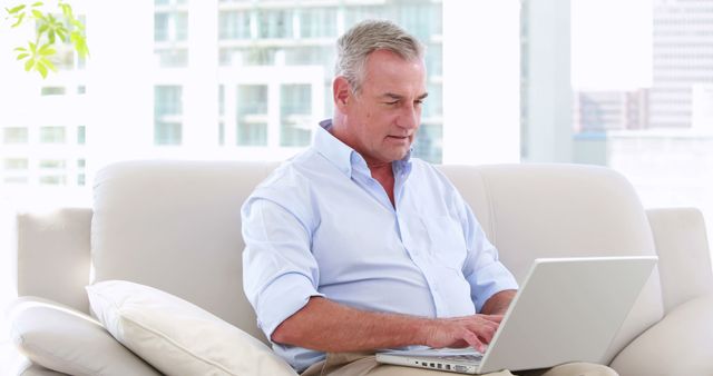 A middle-aged Caucasian man is focused on working on his laptop while comfortably seated on a sofa, with copy space. His casual yet professional demeanor suggests he could be conducting business from home or managing personal affairs online.