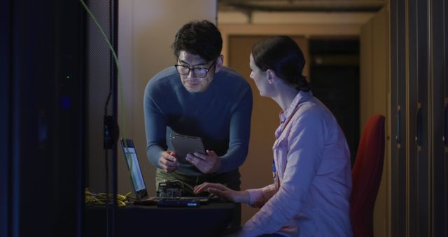 Two IT professionals collaborating in a dimly lit server room, examining solutions on both a tablet and a laptop and managing wire connections. Ideal for use in contexts relating to technology services, data management, IT support, and workplace settings.