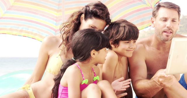 Family of four enjoying beach vacation, sitting close under colorful umbrella, looking at tablet together. Useful for promoting travel destinations, vacation packages, family activities, technology use on holidays.