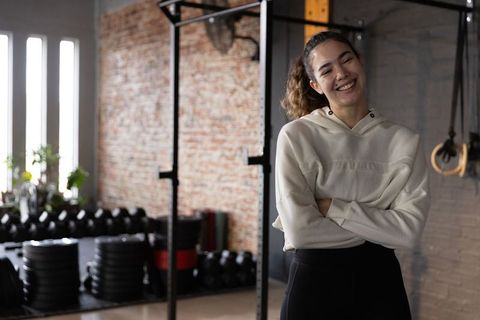 Smiling fit woman standing arms crossed in sunlit gym wearing hoodie and leggings