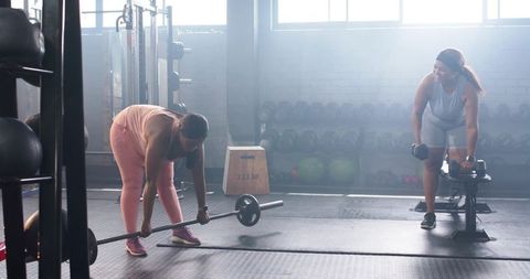 Diverse women doing strength training with barbell and dumbbells in sunlit gym