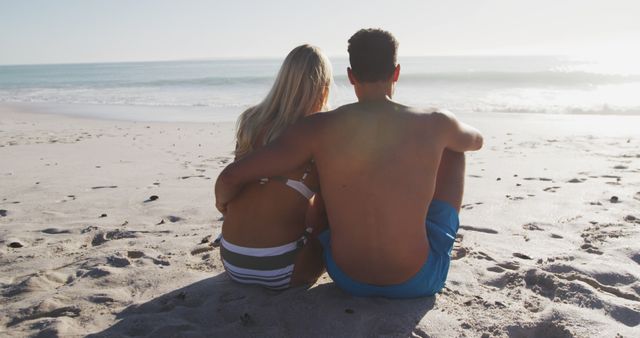 An affectionate couple sits together on a sandy beach, gazing at the ocean during sunrise. This image captures the serene and romantic atmosphere of a peaceful morning by the sea. Perfect for use in travel brochures, romantic getaway advertisements, lifestyle blogs, or articles about relationships and relaxation.