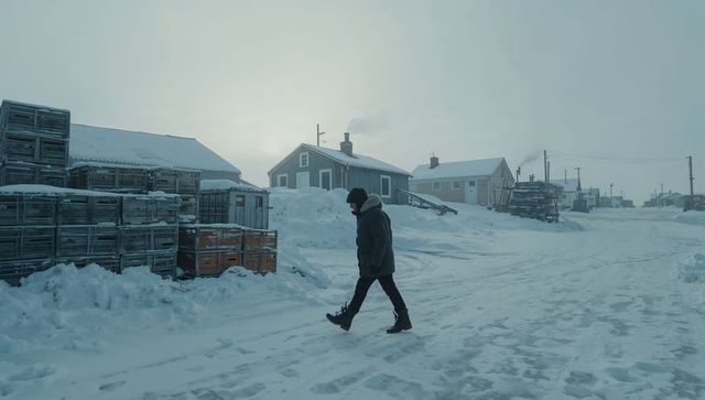 Solitary man walking through snow-covered village wearing heavy parka near stacked crates