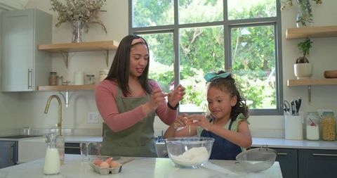 Mother and daughter baking together cracking eggs on marble countertop in farmhouse kitchen