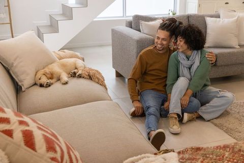 Young couple sitting on living room floor cuddling while golden retriever puppy sleeps on sofa