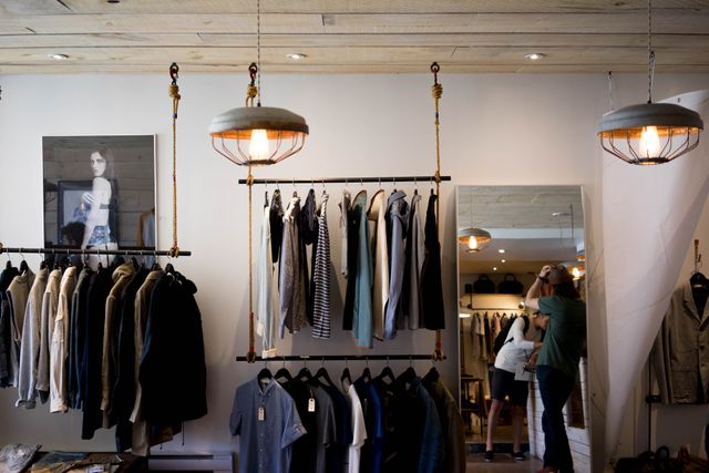Customers exploring a trendy clothing store with an array of modern apparel hanging on racks. Warm, stylish hanging lights create a cozy atmosphere, adding to the boutique’s modern vibe. Perfect for depicting fashion retail environments, contemporary interior design, and shopping experiences.