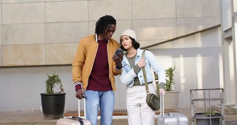 Diverse couple checking smartphone while pulling wheeled suitcases outside modern building