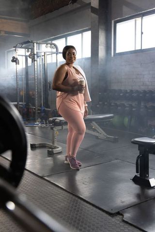 African american woman in pink activewear standing in gym holding shaker and towel