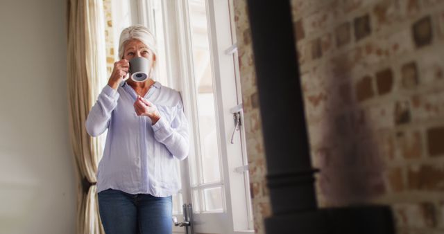 Happy senior caucasian woman standing in bedroom, looking through window and drinking coffee - Download Free Stock Photos Pikwizard.com