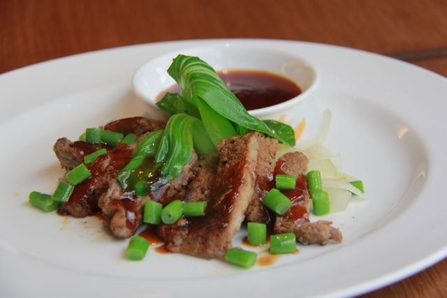 Grilled beef slices, topped with chopped green onions, paired with bok choy and a flavorful dipping sauce on a white plate. Ideal for use in culinary websites, food blogs, or restaurant menus to showcase gourmet dining, healthy meal options, or Asian-inspired dishes.